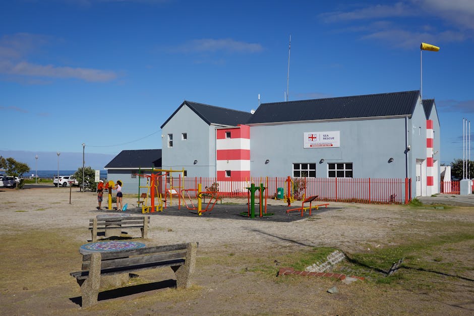 The image features a light blue building with red and white striped accents, labeled as a Sea Rescue station, situated under a clear blue sky. In front of the building, there is a small playground wit