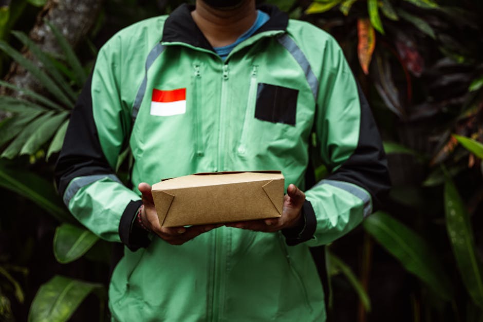 Close-up of a delivery person wearing a green uniform with a flag patch and black accents, holding a rectangular brown cardboard parcel with folded edges. The background features lush green foliage, i