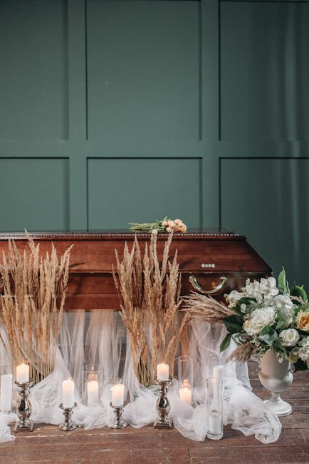 An indoor floral display featuring a wooden casket draped with sheer white fabric, surrounded by tall, beige dried grasses and several small lit candles in silver holders. To the right, there is a whi