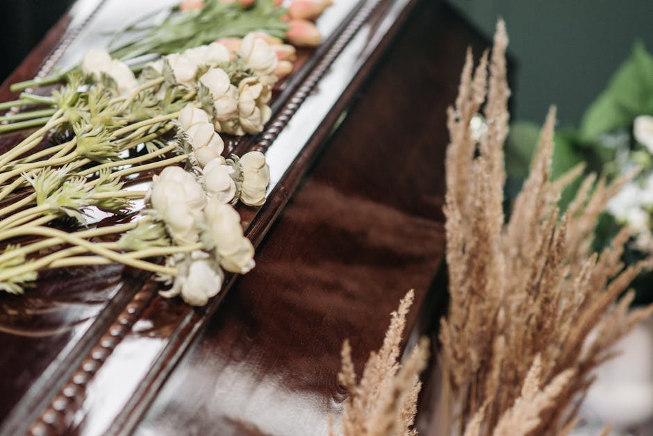 A close-up view of a floral tribute arranged on top of a polished wooden surface, featuring a bouquet of white roses and delicate peach-colored tulips with green stems and leaves, wrapped with subtle 