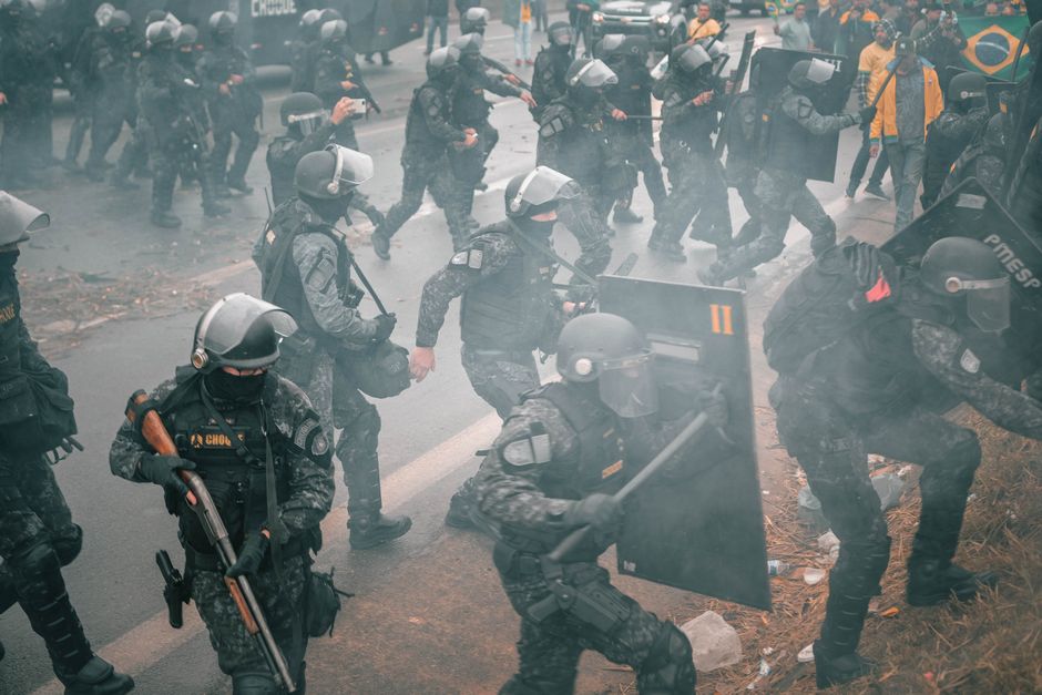 The image depicts a group of riot police officers in tactical gear, including helmets and body armor, advancing on a street with a misty, tense atmosphere. Some officers are holding shields, while oth
